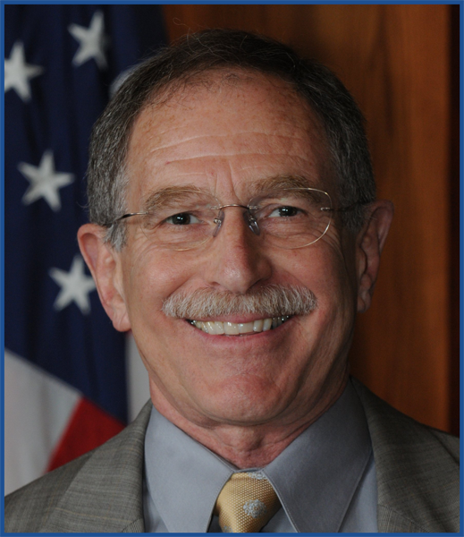 Headshot of a man with gray hair, a moustache, and glasses, wearing a gray blazer and tie, with the corner of an American flag visible in the background.