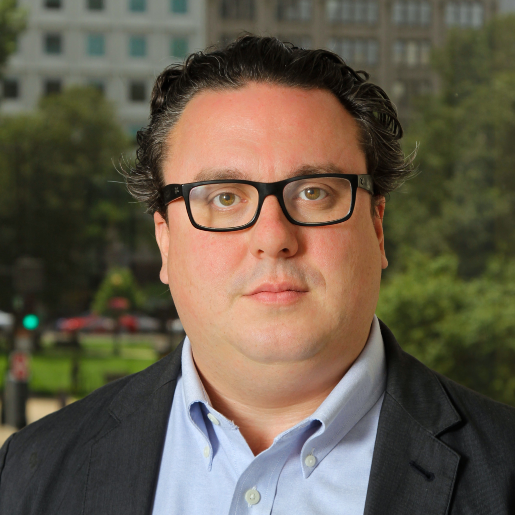 Headshot of a brunet man wearing glasses, a blue collared shirt, and a gray blazer.