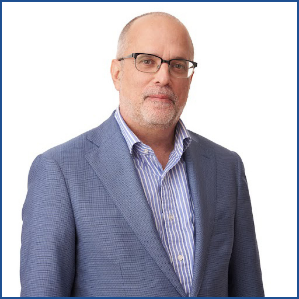 Headshot of a man with receding salt & pepper hair, a beard, and horn-rimmed glasses wearing a light blue blazer and shirt.