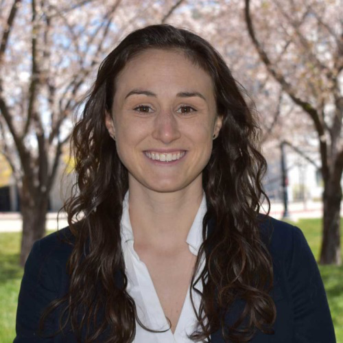 Headshot of a woman with back-length wavy brown hair standing outside and wearing a navy blazer with a white collared shirt.