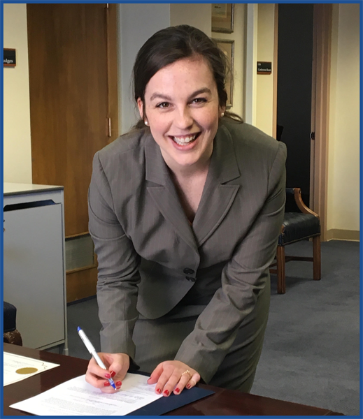 A woman in a gray suit standing at a desk and smiling at the camera while signing a paper.