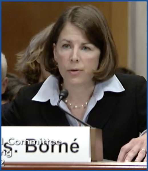 Headshot of a brunette woman wearing a bob, black blazer, a white collared shirt, and a short pearl necklace speaking into a microphone.