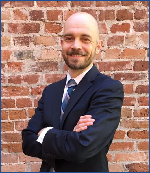 Headshot of a bearded bald man in a navy suit standing in front of a brick wall